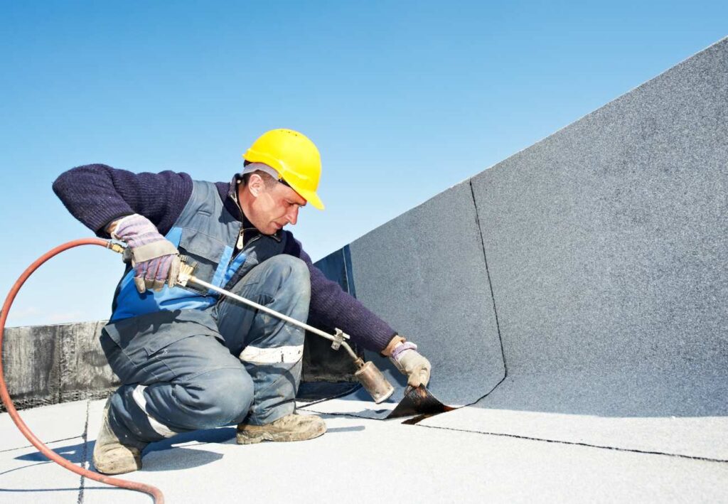 A worker in a yellow helmet is applying adhesive to a flat roof with a brush.