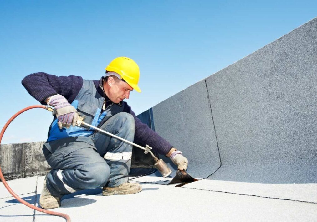 A worker in a yellow helmet is applying adhesive to a flat roof with a brush.