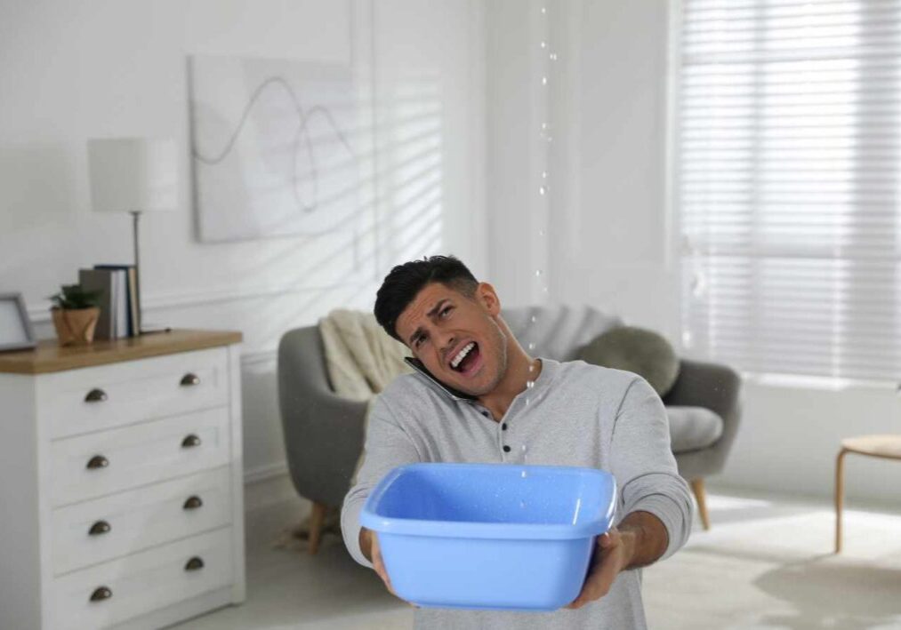 A man is indoors holding a blue basin to catch water leaking from the ceiling.