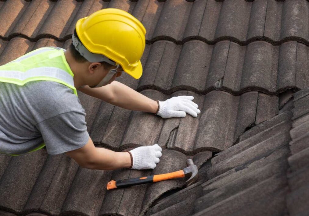 A person in safety gear is inspecting or repairing a tiled roof with a hammer in hand.