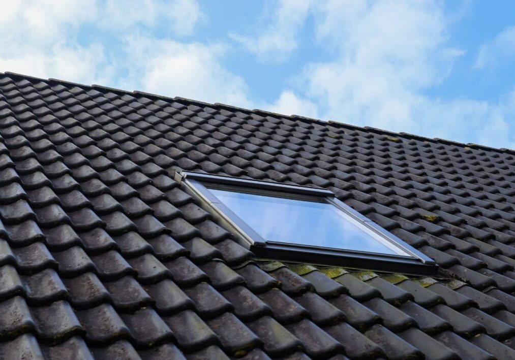 A skylight window installed on a sloped roof with dark tiles against a clear blue sky.